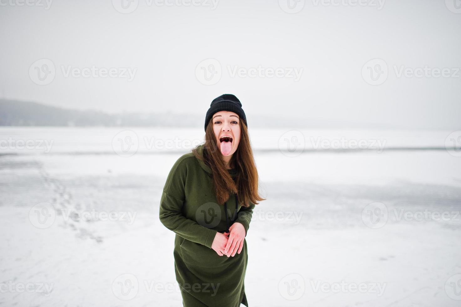 garota engraçada usa moletom verde longo, jeans e chapéu preto mostra uma língua no lago congelado em dia de inverno. foto