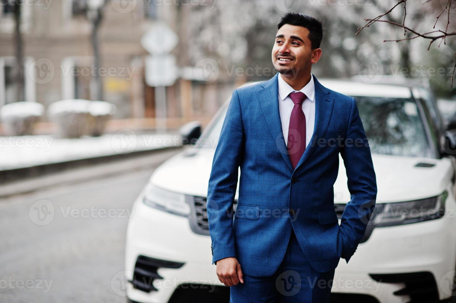 modelo de homem elegante indiano elegante terno posado em dia de inverno contra carro suv de negócios branco. foto