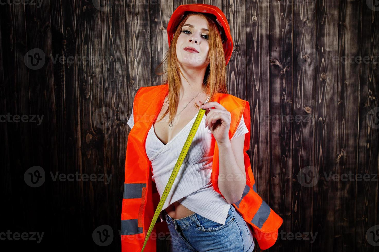 mulher engenheira em laranja proteger o capacete e a jaqueta de construção contra o fundo de madeira, segurando a fita métrica. foto