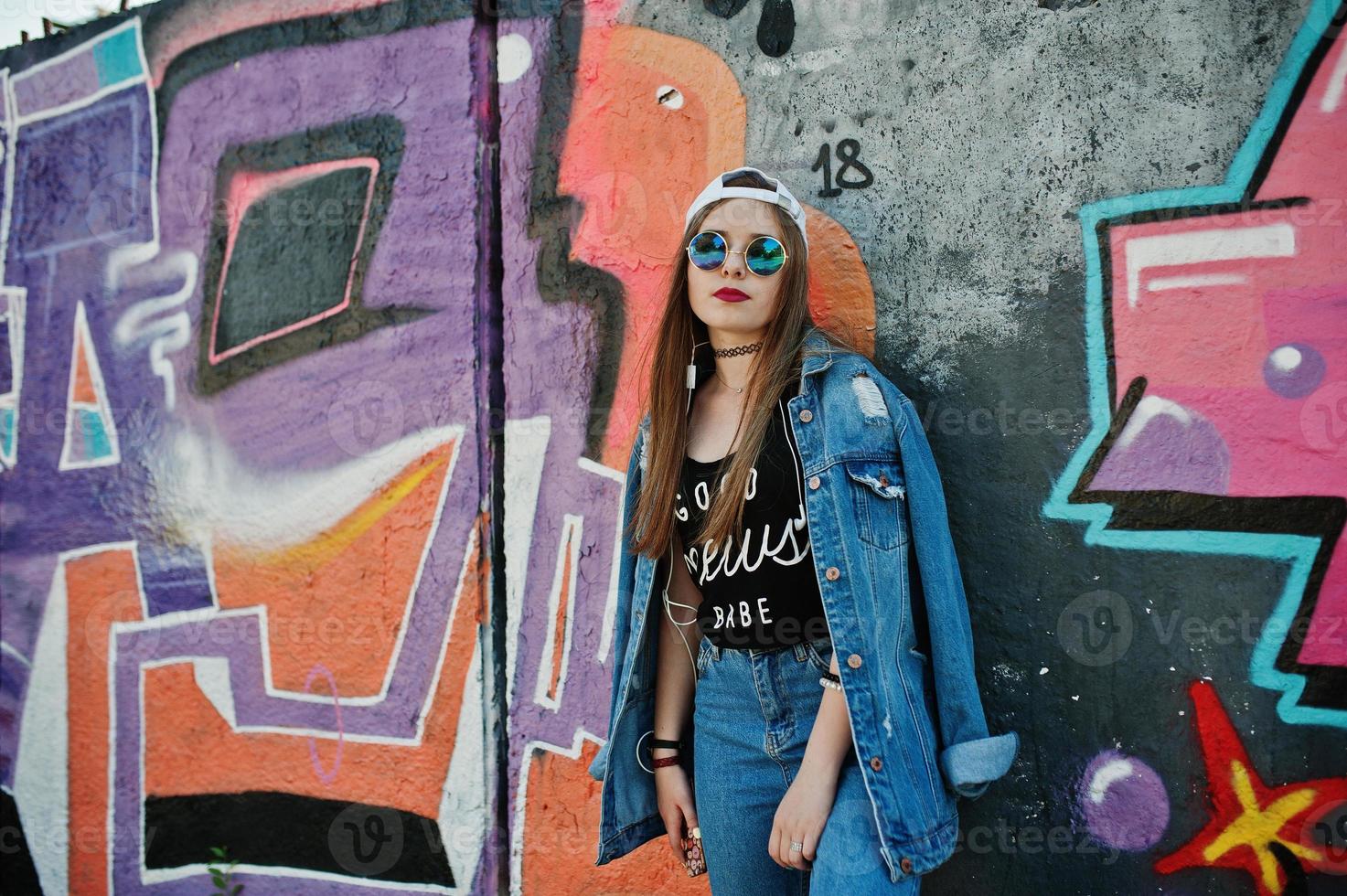 menina elegante casual hipster no boné, óculos escuros e calça jeans, ouvindo música de fones de ouvido do telefone celular contra grande parede de graffiti. foto