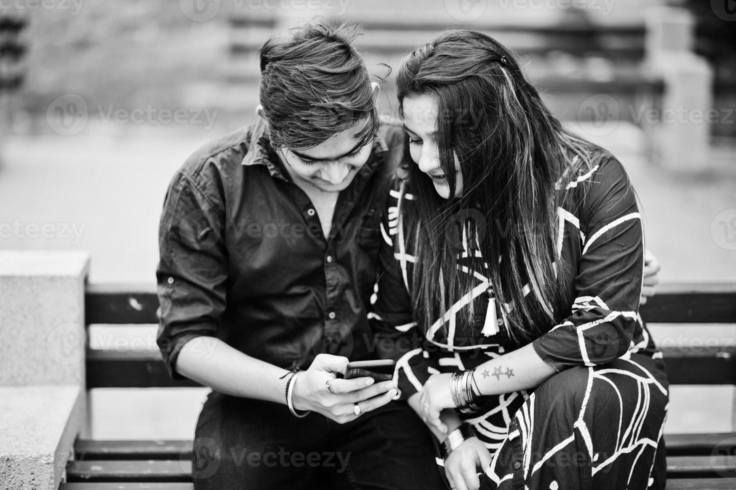 casal indiano posou ao ar livre, sentados no banco juntos e olhando para o celular. foto