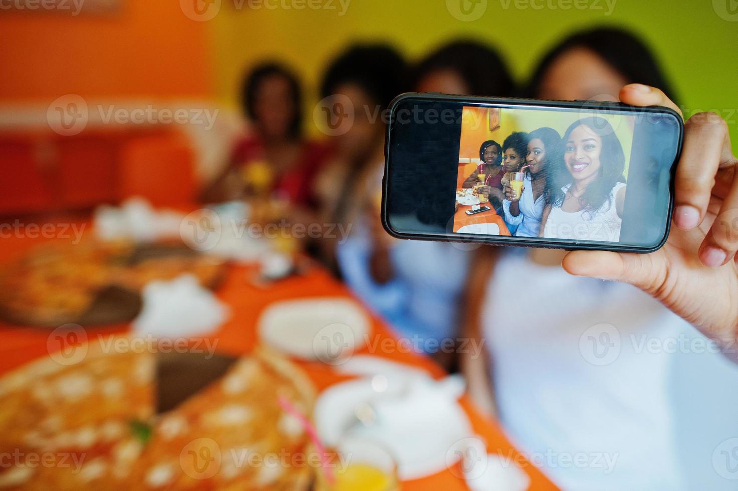 feche a tela do telefone da foto de quatro jovens africanas no restaurante colorido brilhante comendo pizza, se divertindo juntos e fazendo selfie.