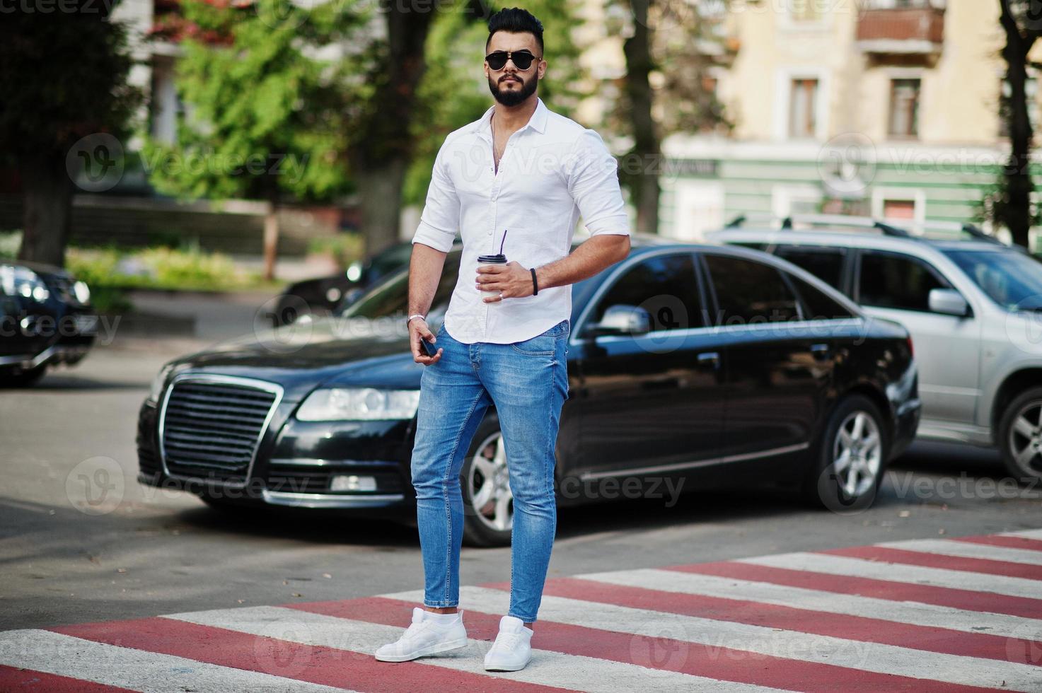 elegante modelo de homem árabe alto na camisa branca, jeans e óculos de sol posou na rua da cidade. barba cara árabe atraente com café contra carro de negócios preto. foto