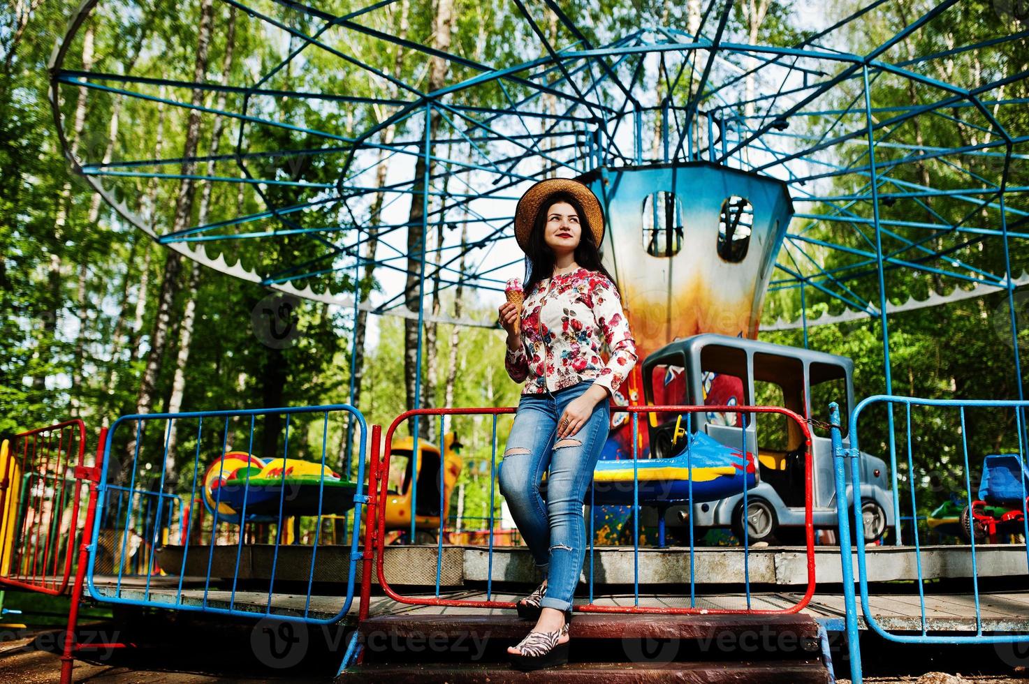 retrato de menina morena de óculos cor de rosa e chapéu com sorvete no parque de diversões. foto