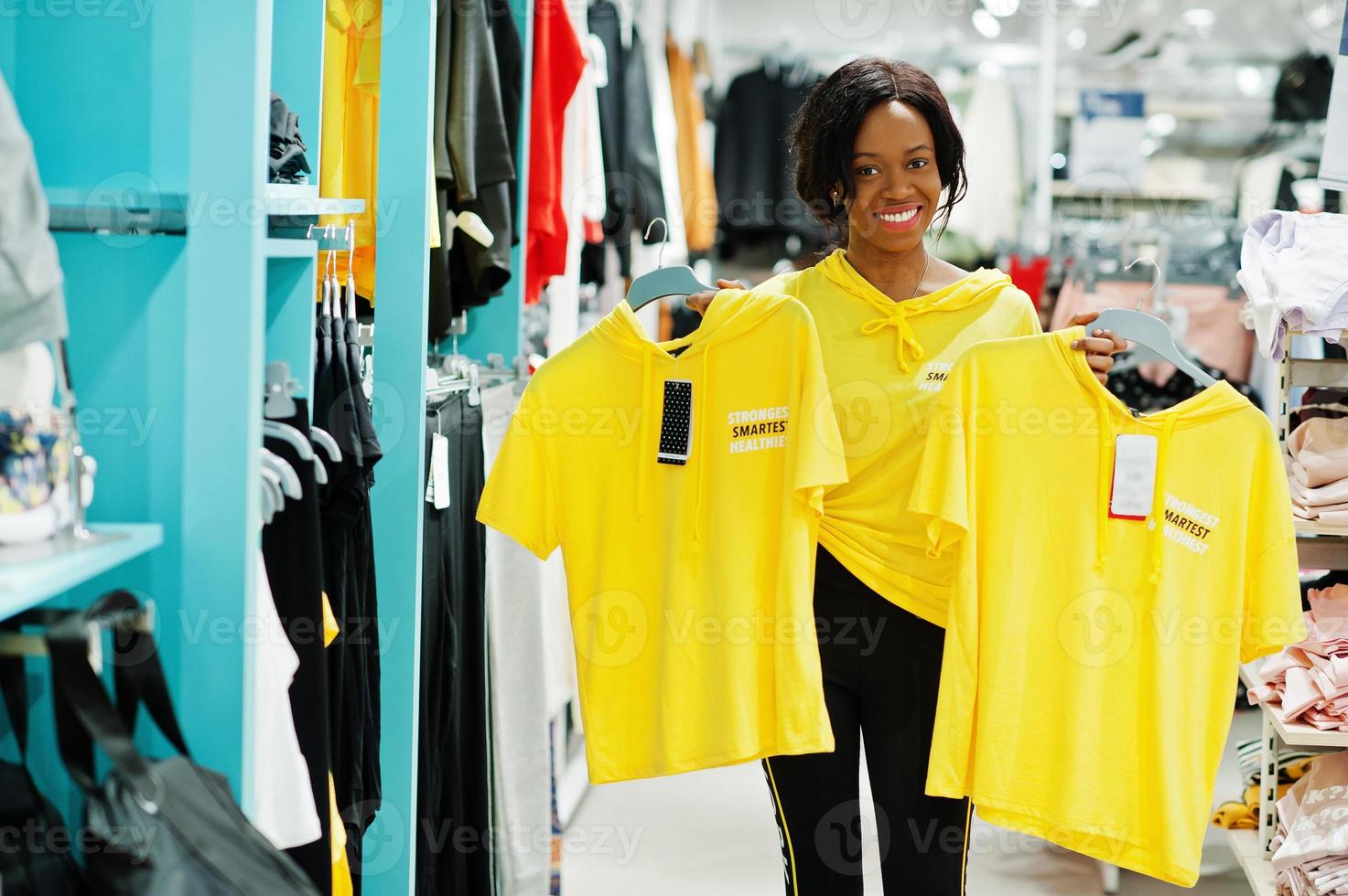 mulheres afro-americanas em agasalhos fazendo compras no shopping de roupas esportivas contra prateleiras. ela escolhe camiseta amarela. tema da loja de esportes. foto