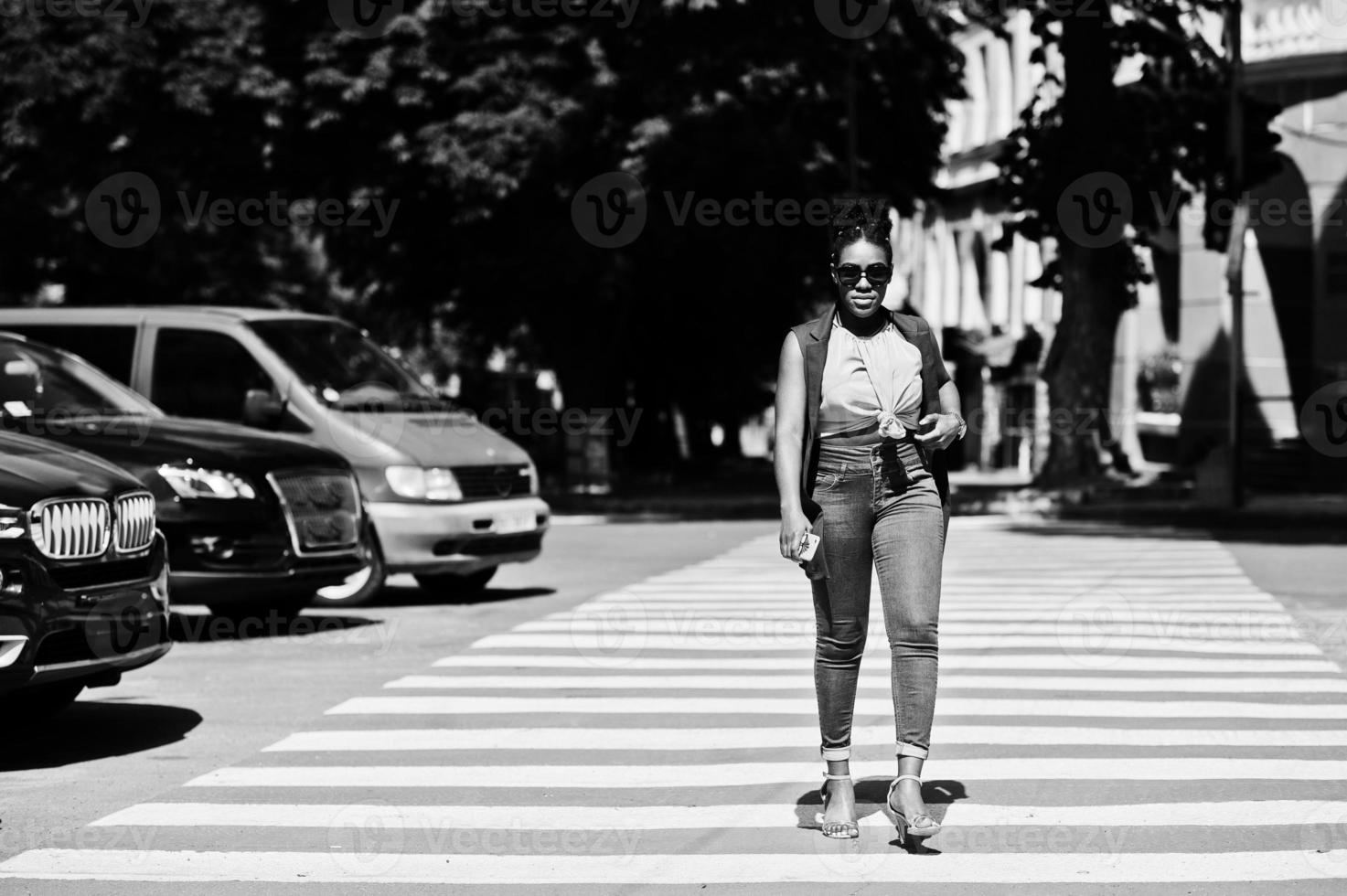 elegante mulher afro-americana andando na faixa de pedestres ou travessia de pedestres com o celular na mão. foto