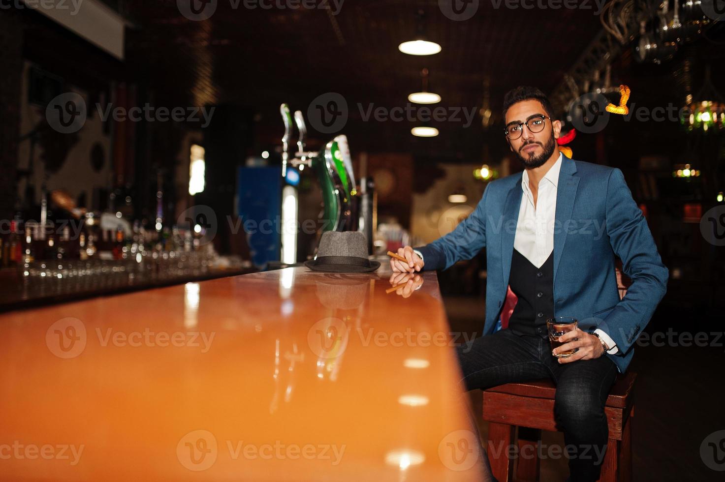 homem árabe bem vestido bonito com copo de uísque e charuto posou no pub. foto