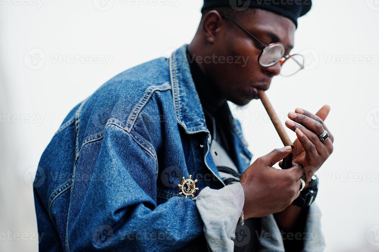 homem afro-americano de jaqueta jeans, boina e óculos, acende um charuto. foto