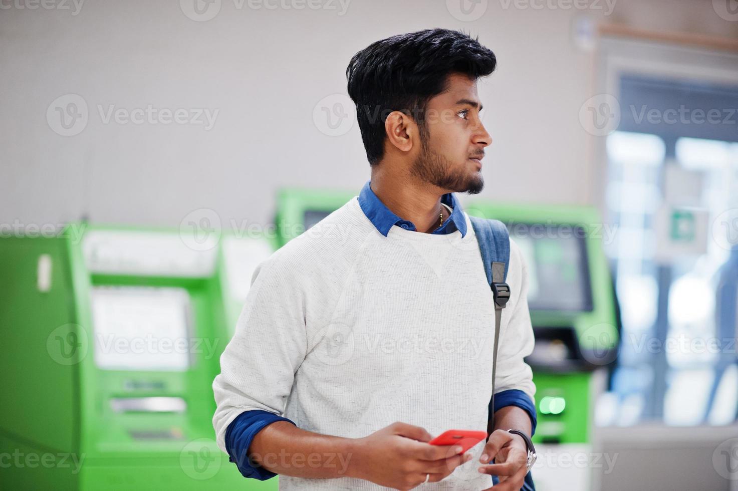 jovem asiático elegante com celular e mochila contra a linha de atm verde. foto