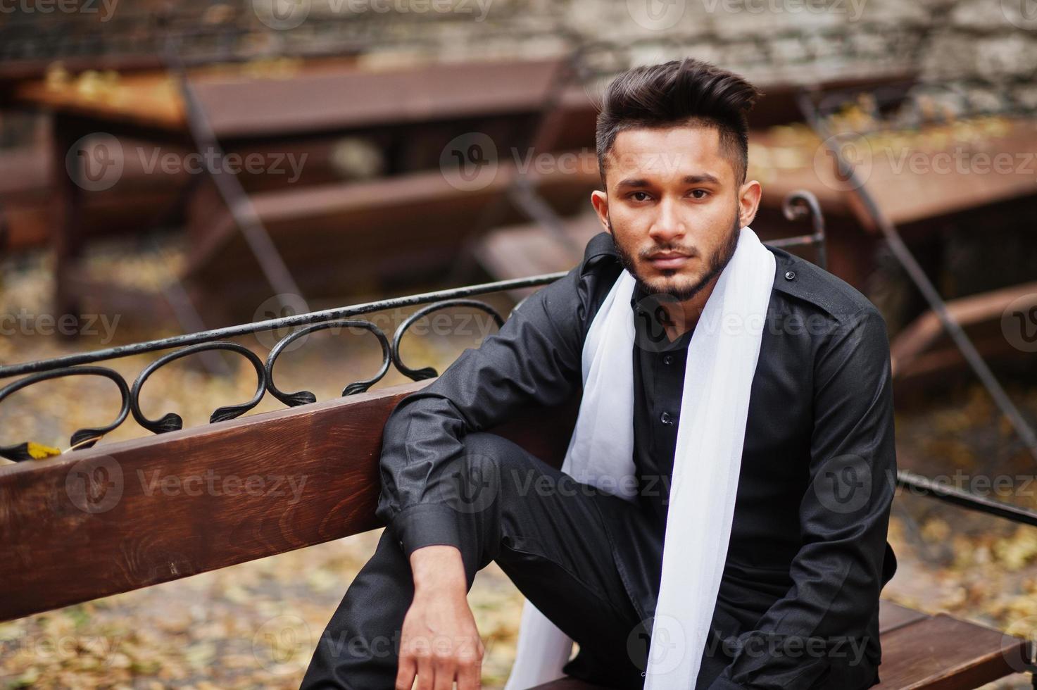 homem elegante indiano em roupas tradicionais pretas com lenço branco posou ao ar livre sentado no banco. foto