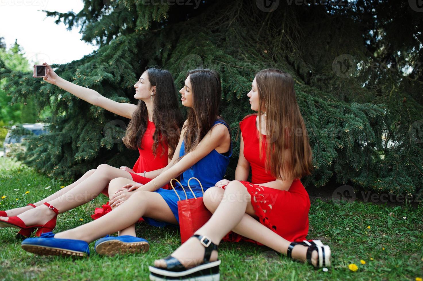 menina de três adolescentes em vestidos azuis e vermelhos posou ao ar livre e fazendo selfie no celular. foto