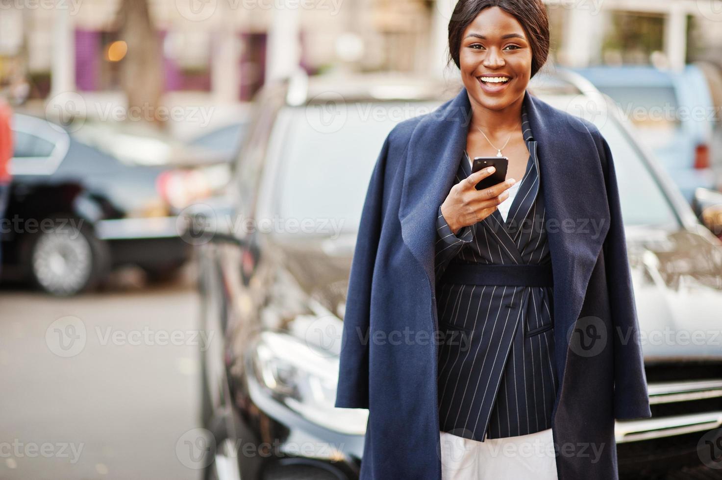 sucesso elegante mulher afro-americana de casaco contra carro suv de negócios preto olhando para o telefone. foto