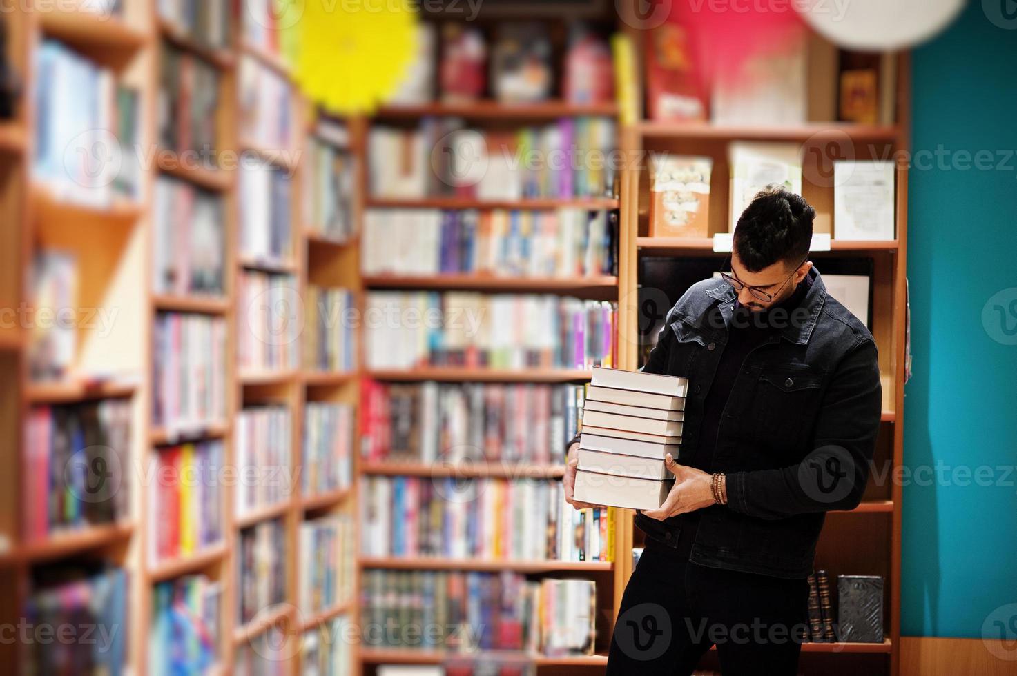 homem alto estudante árabe inteligente, use jaqueta jeans preta e óculos, na biblioteca com pilha de livros. foto