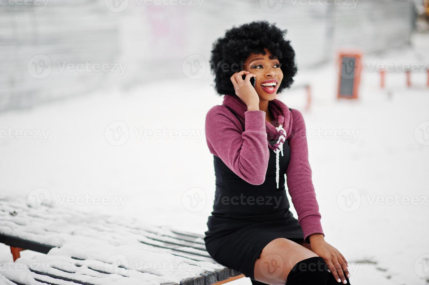 mulher afro-americana de cabelo encaracolado posou no dia de inverno, sentado no banco e falando no celular. foto