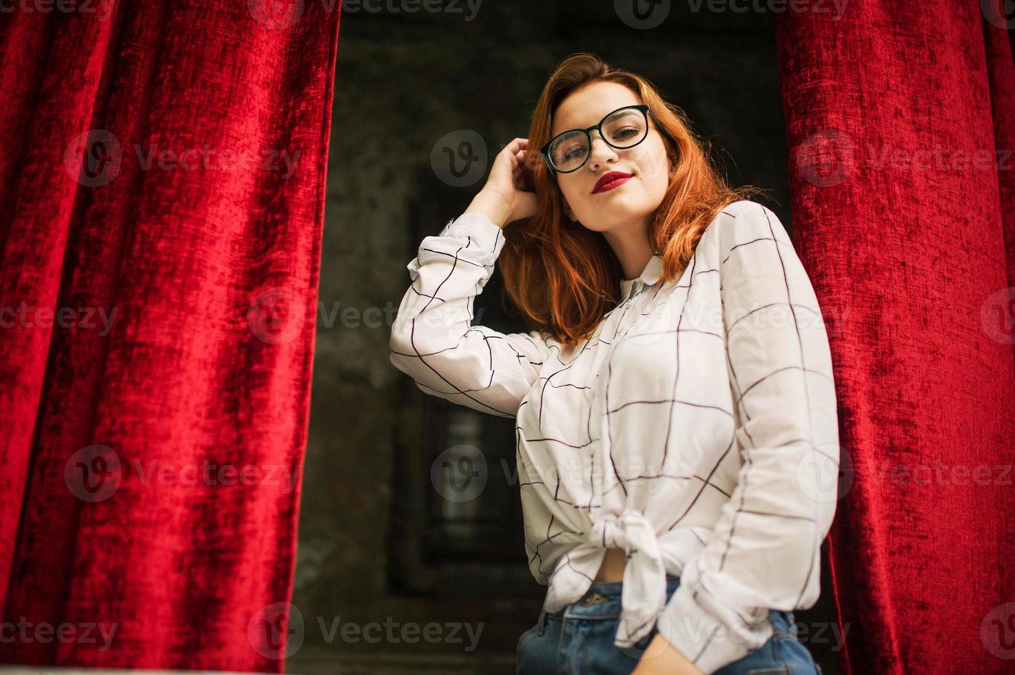 mulher ruiva atraente em óculos, use blusa branca posando no arco de cortinas vermelhas abertas. foto