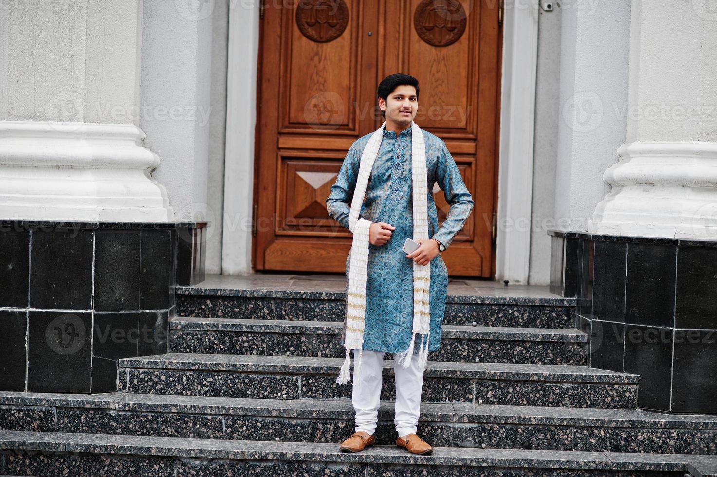 homem indiano usa roupas tradicionais com lenço branco posou ao ar livre contra a porta do prédio com o celular na mão. foto