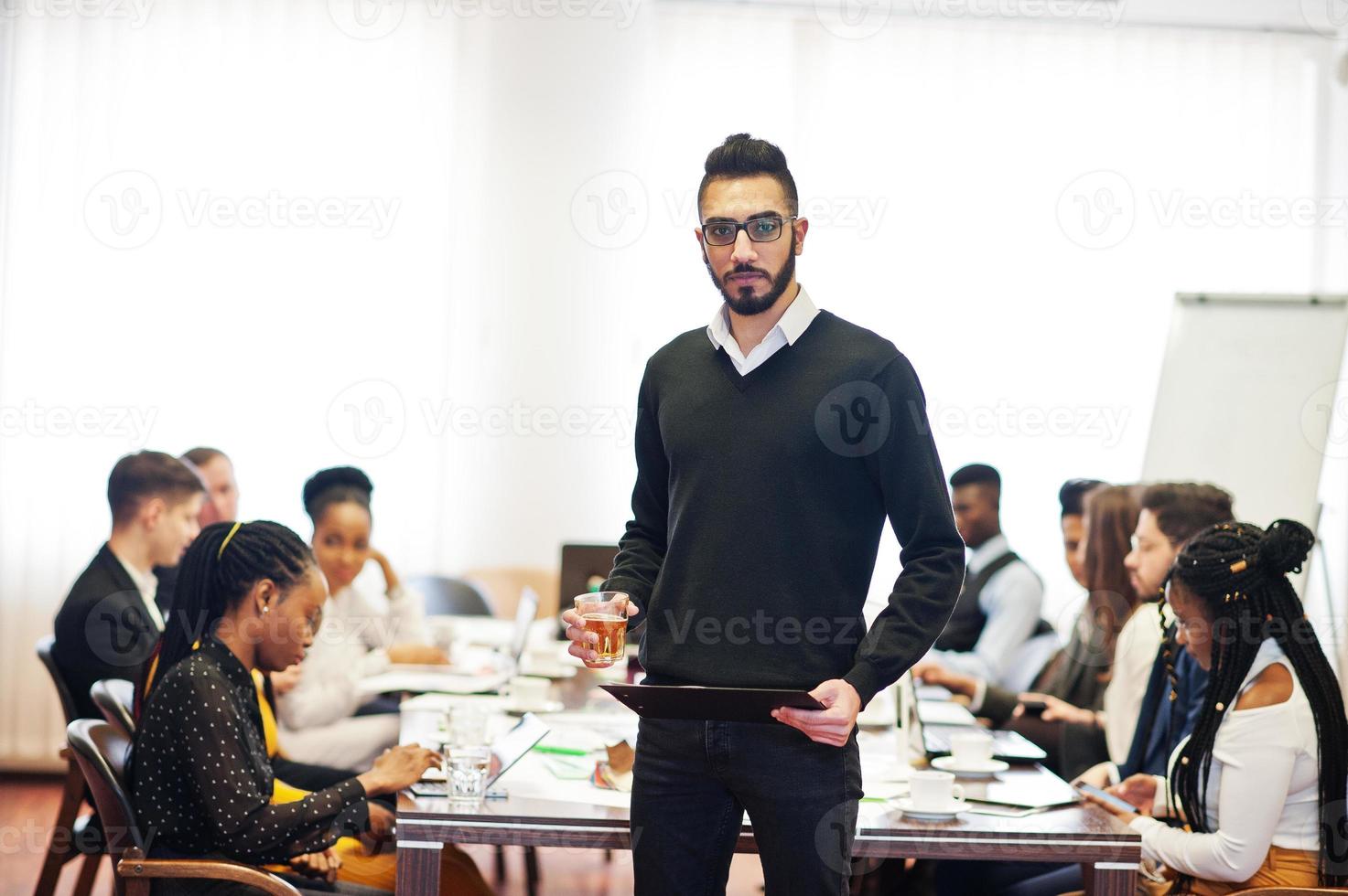rosto de homem de negócios árabe bonito, segurando a prancheta e copos de bebida no fundo da reunião de equipe multirracial de pessoas de negócios, sentado na mesa do escritório. foto