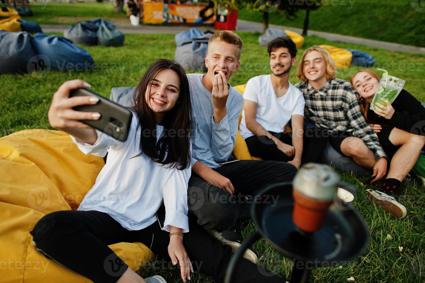 jovem grupo multiétnico de pessoas assistindo filme no poof no cinema ao ar livre e fazendo selfie no telefone. foto