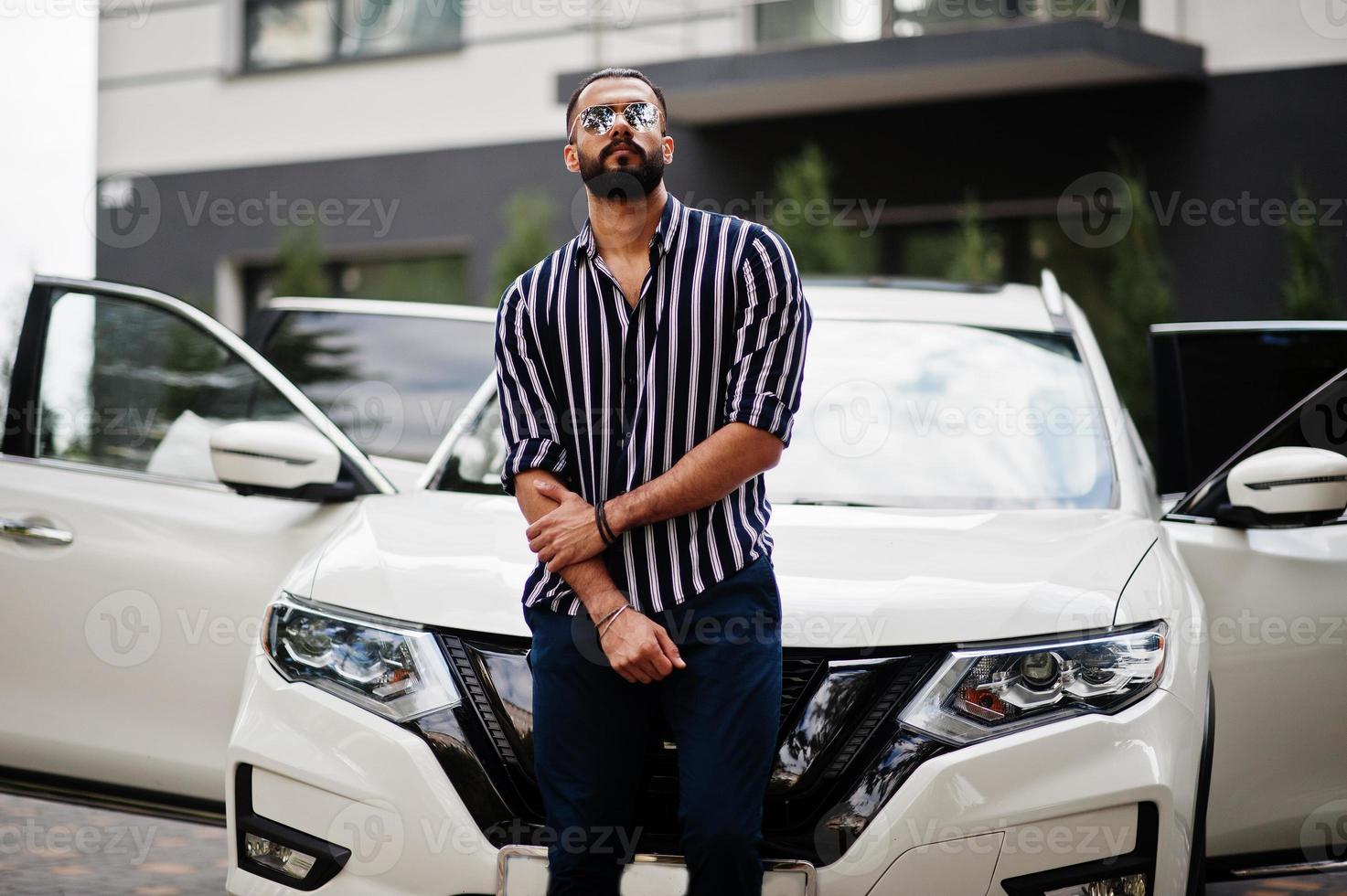 homem árabe bem sucedido usa camisa listrada e óculos de sol posam perto de seu carro suv branco. homens árabes elegantes no transporte. foto