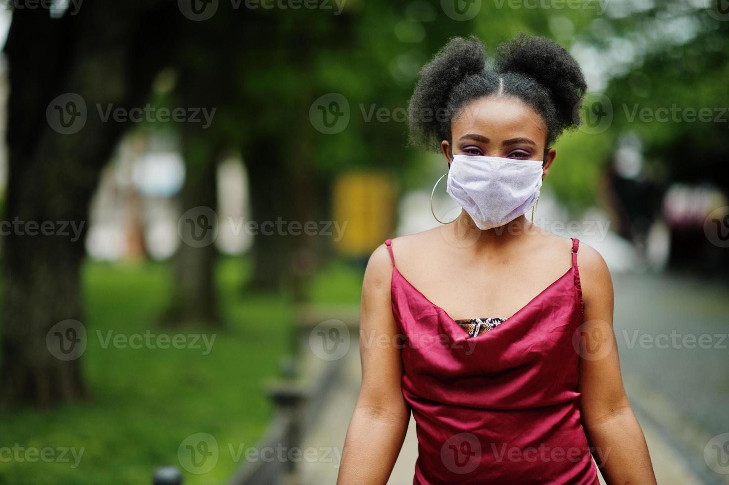 covid-19, vírus infeccioso. mulher africana com cabelos cacheados, usa vestido de seda vermelho e máscara médica descartável, se preocupa com sua saúde e protege em situações perigosas. foto