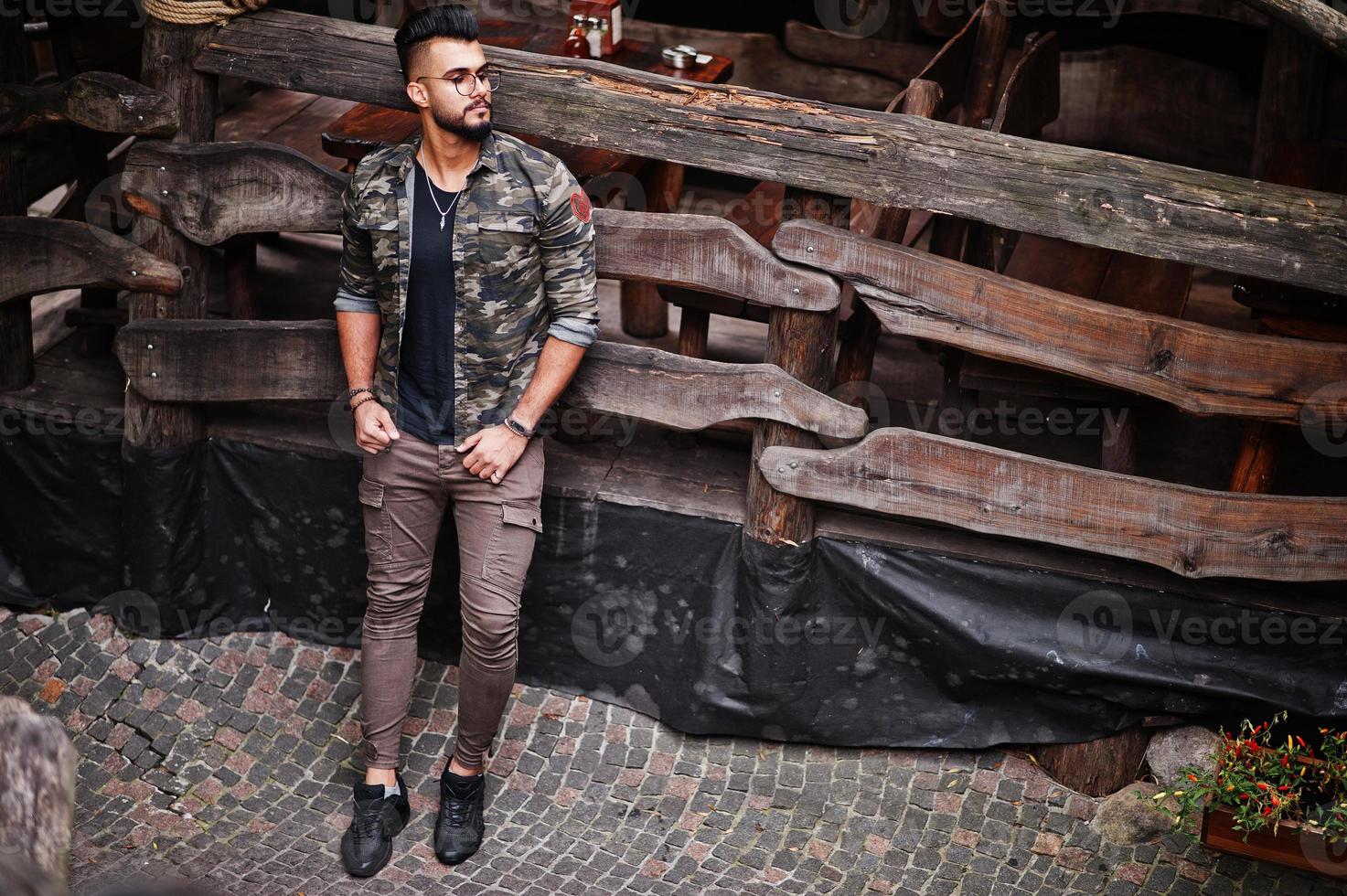 incrível lindo homem macho de barba árabe alta de óculos e jaqueta militar posou ao ar livre contra a casa rústica de madeira. foto