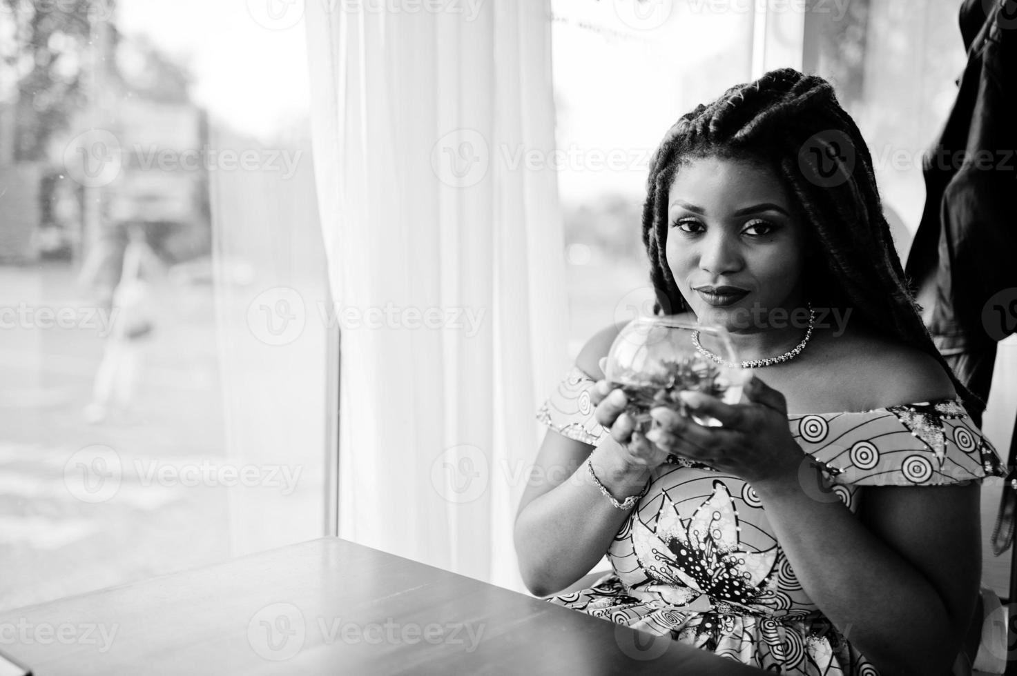 linda garota afro-americana de pequena altura com dreadlocks, use vestido amarelo colorido, sentado no café com flor no vidro. Preto e branco. foto