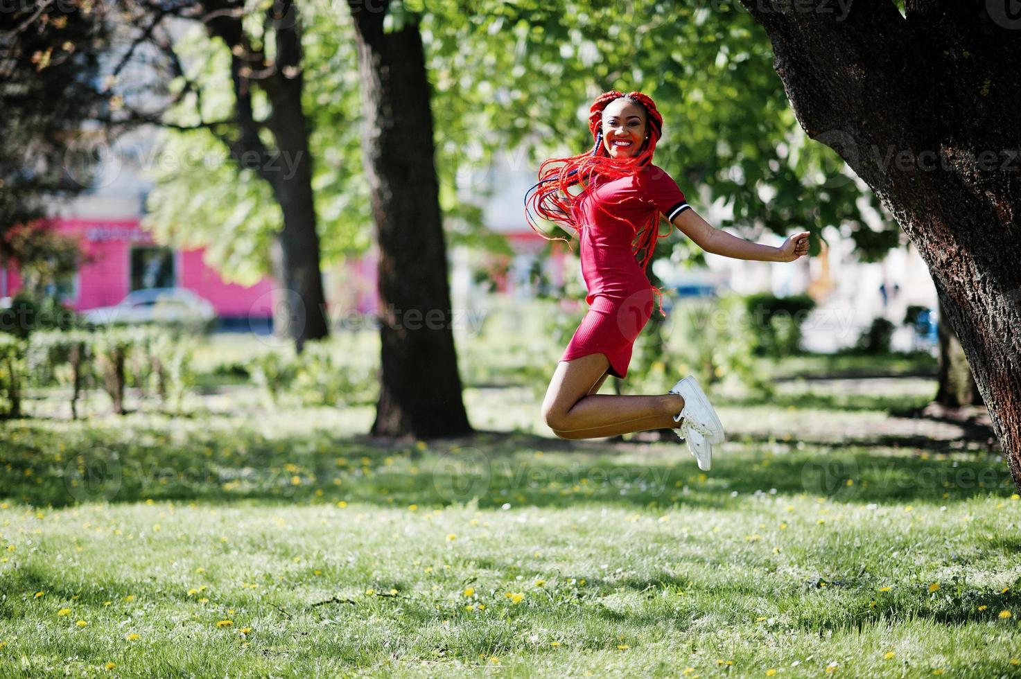 linda e magra garota afro-americana de vestido vermelho com dreadlocks salta ao ar livre no parque primavera. elegante modelo preto. foto