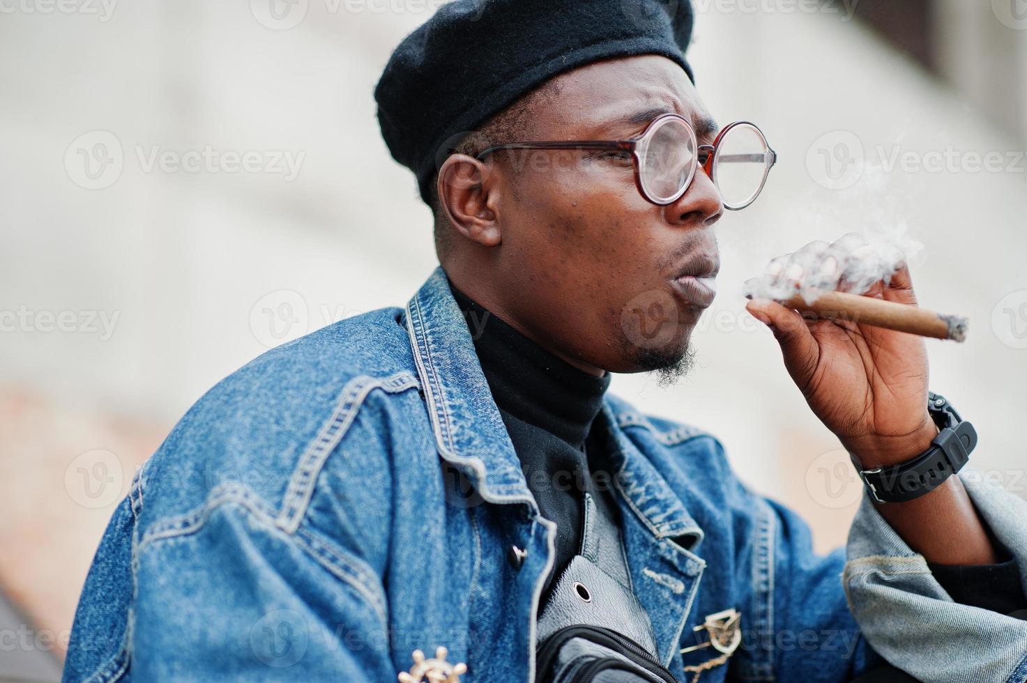 feche o retrato do homem afro-americano na jaqueta jeans, boina e óculos, fumando charuto. foto