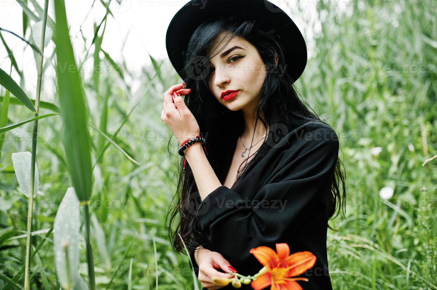 garota sensual toda em preto, lábios vermelhos e chapéu. mulher dramática gótica segura flor de lírio laranja na cana comum. foto