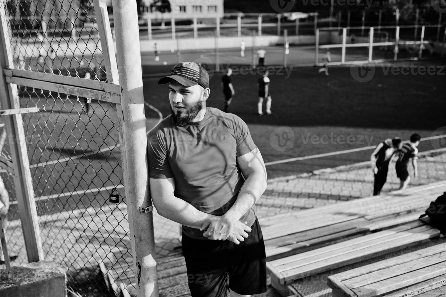 jovem homem musculoso barbudo brutal usar camisa vermelha, shorts e boné no estádio. foto