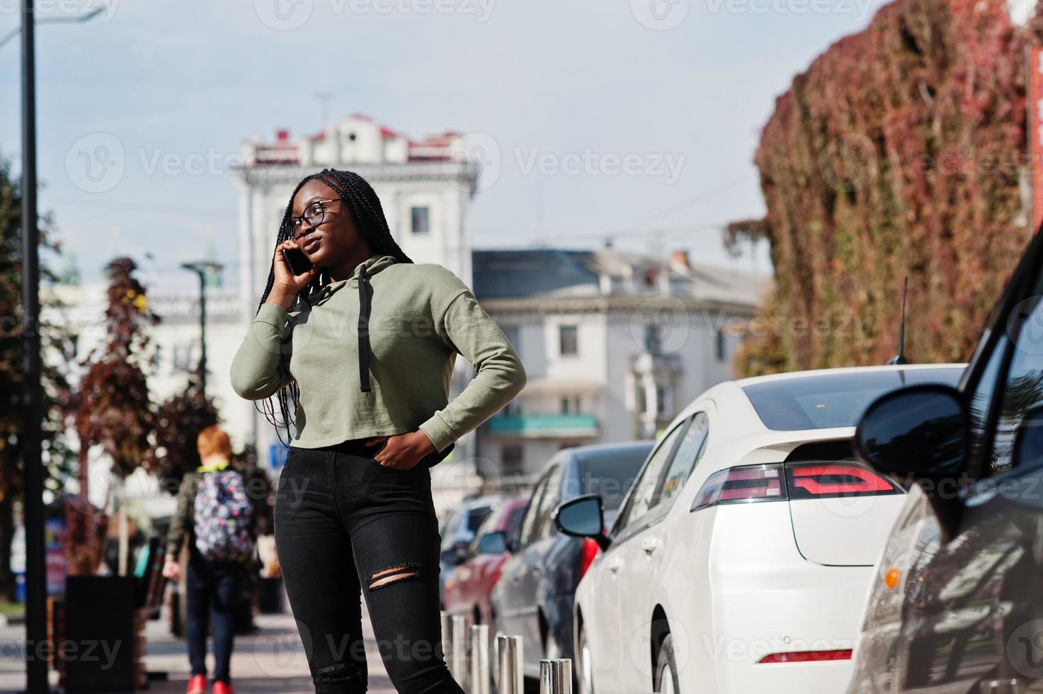 retrato da cidade de uma jovem fêmea de pele escura positiva vestindo capuz verde e óculos andando no estacionamento com telefone celular. foto