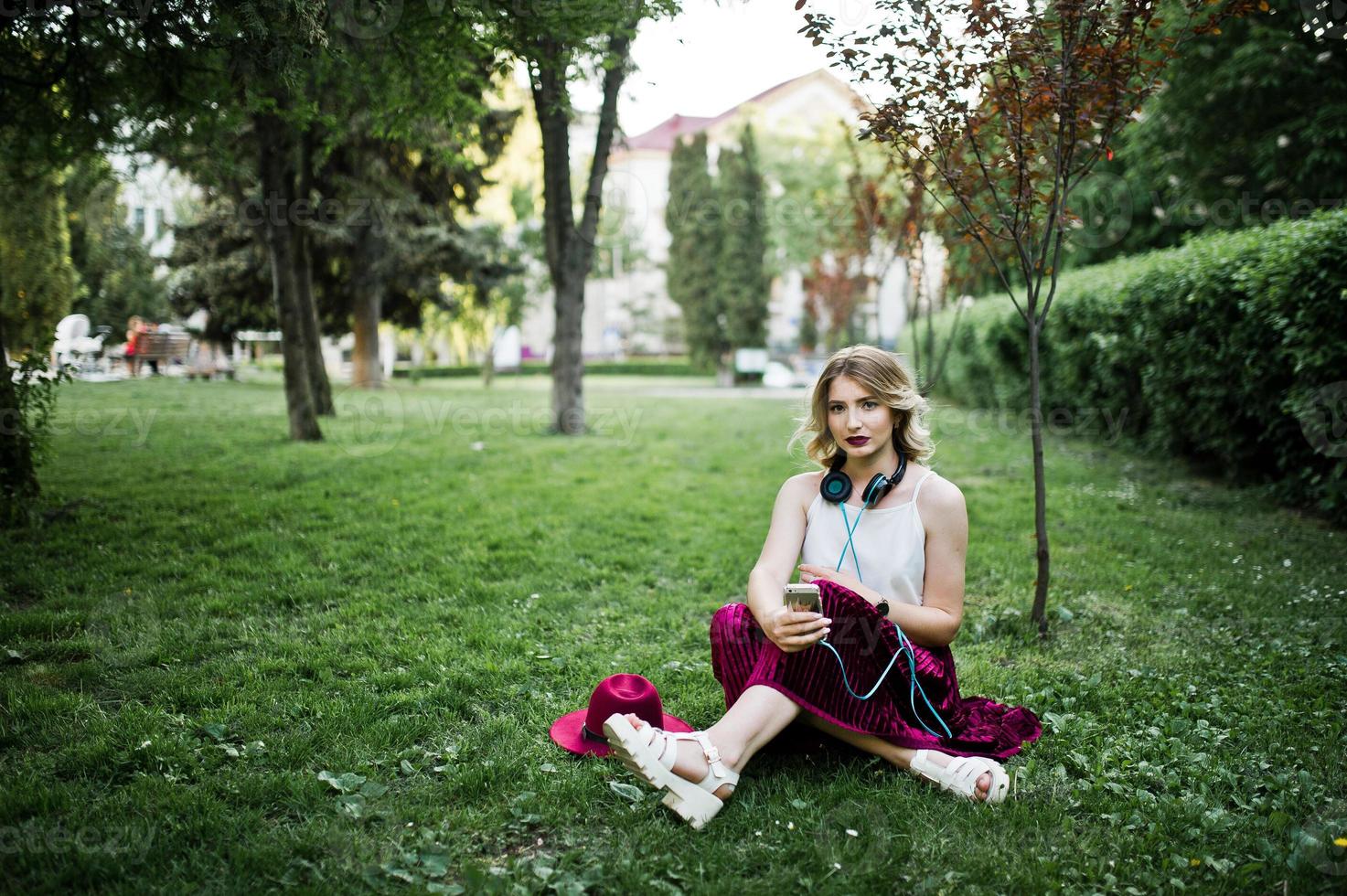 menina modelo loira elegante e bonita na saia de veludo vermelho elegante, blusa branca e chapéu, sentado na grama verde no parque com telefone e fones de ouvido. foto