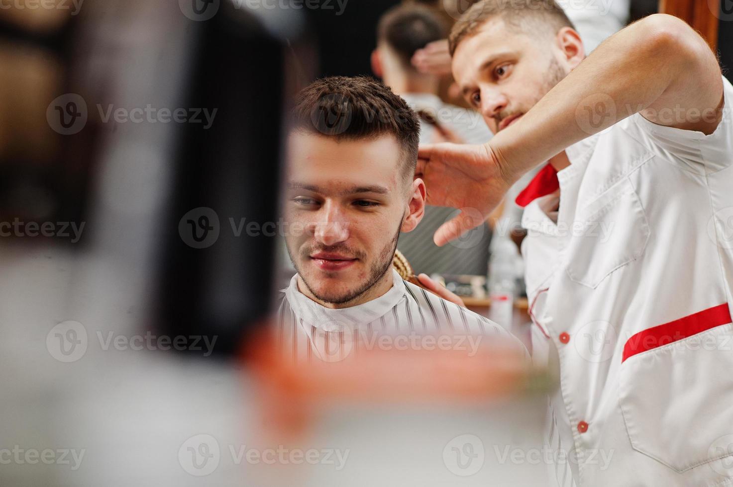 jovem barbudo cortando o cabelo pelo cabeleireiro enquanto está sentado na cadeira na barbearia. alma de barbeiro. foto