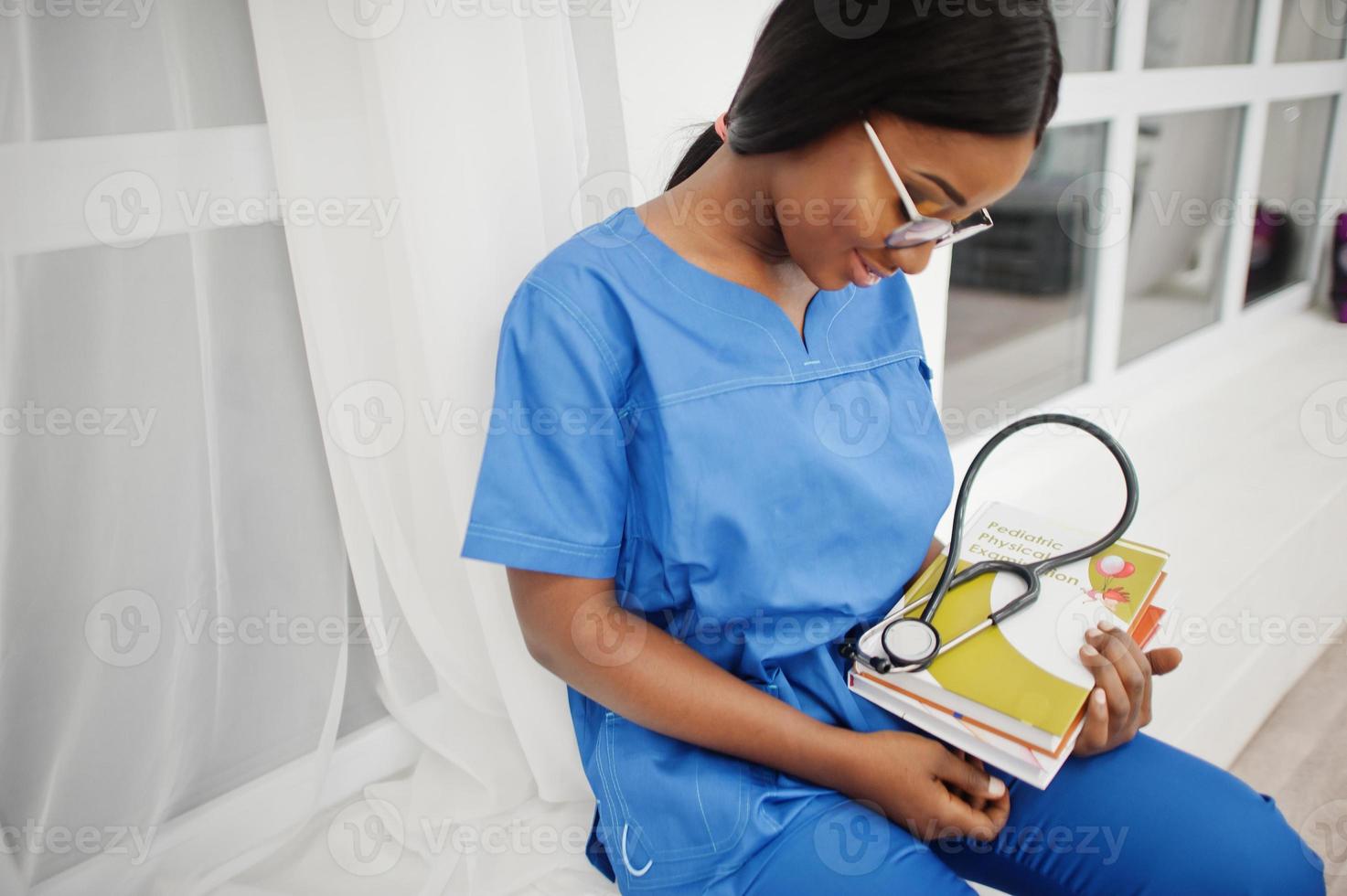 retrato de feliz feminino americano africano jovem médico pediatra no casaco uniforme azul e estetoscópio com livros nas mãos. saúde, médico, especialista em medicina - conceito. foto