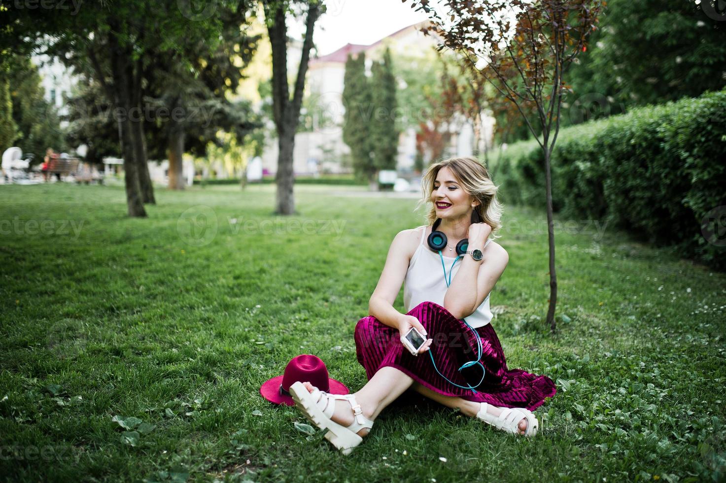 menina modelo loira elegante e bonita na saia de veludo vermelho elegante, blusa branca e chapéu, sentado na grama verde no parque com telefone e fones de ouvido. foto