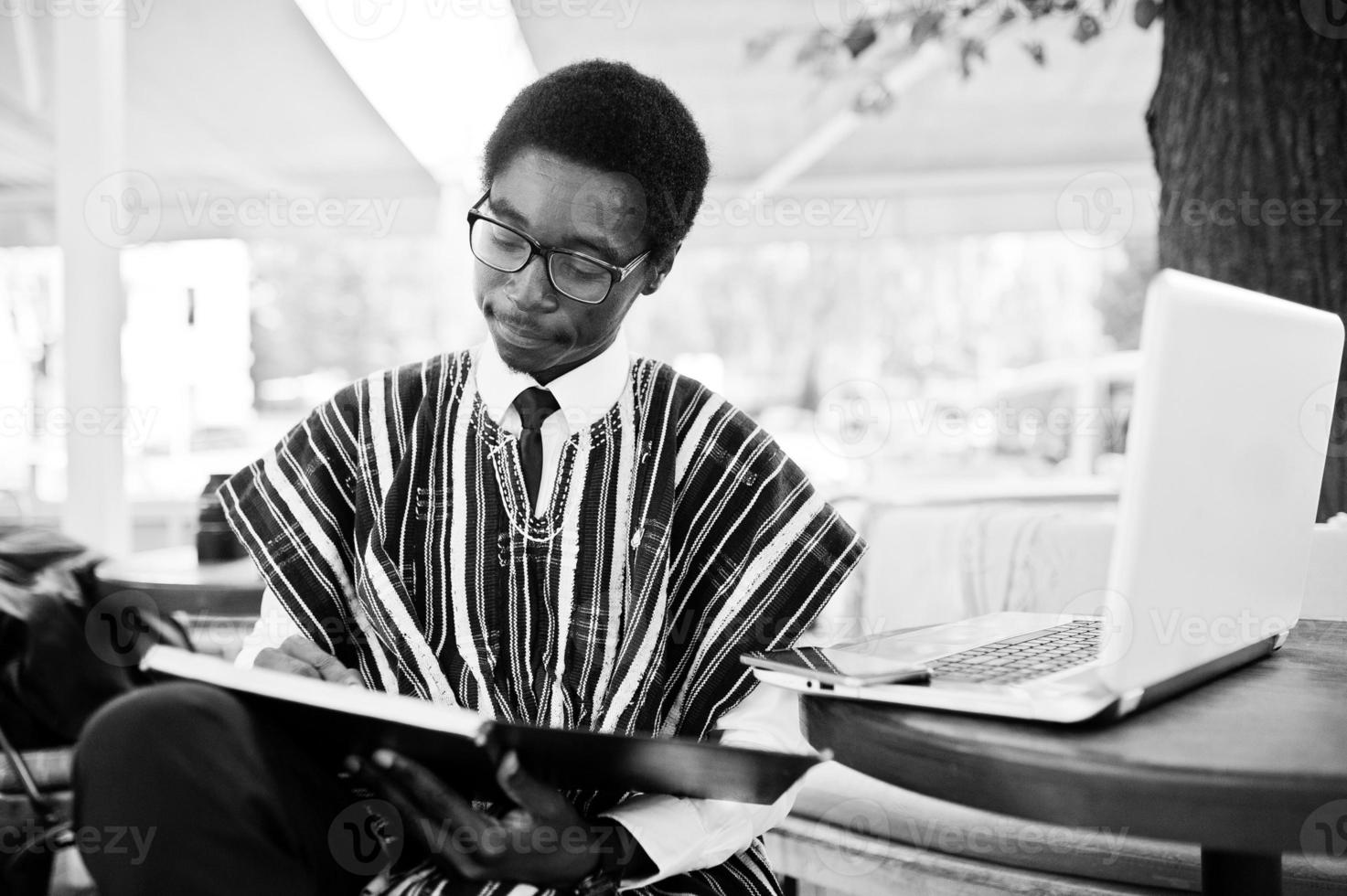 homem africano em roupas tradicionais e óculos sentado atrás do laptop no caffe ao ar livre e olhando em seu notebook. foto