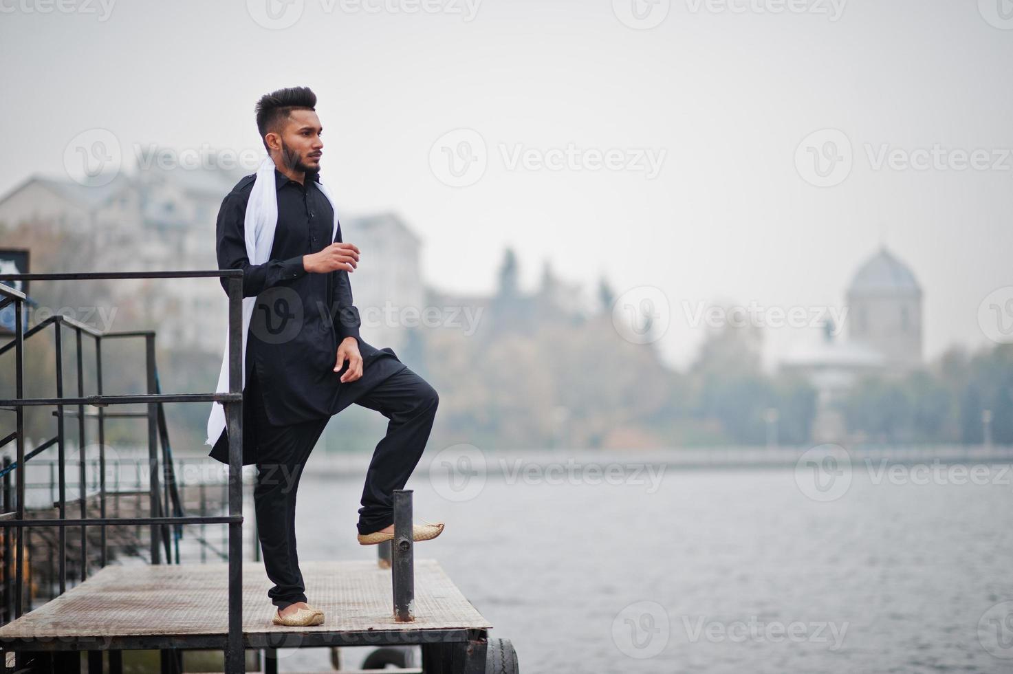 homem elegante indiano em roupas tradicionais pretas com lenço branco posou ao ar livre contra cais nebuloso do mar. foto