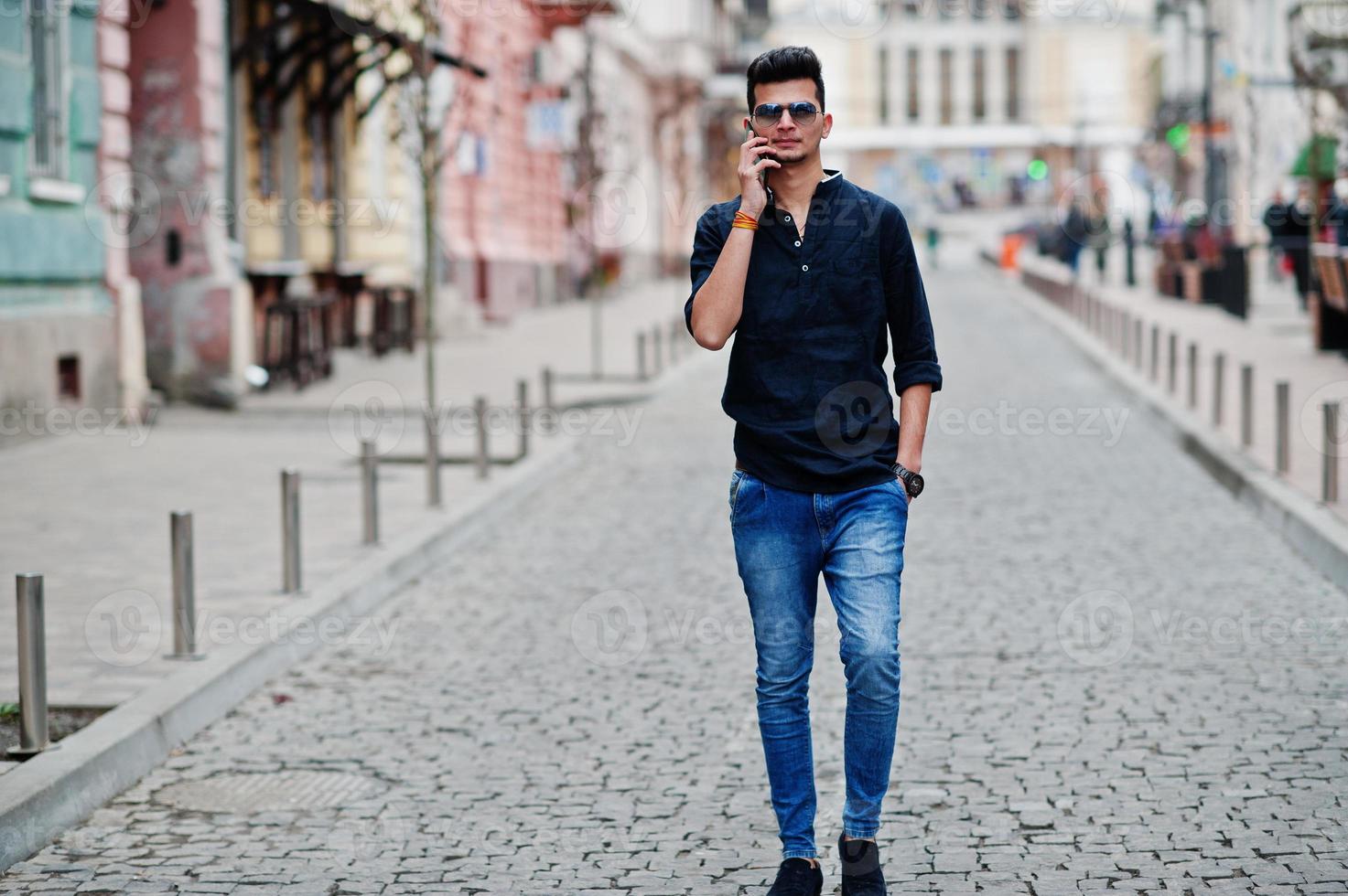 homem elegante modelo indiano em roupas casuais, camisa preta e óculos de sol posou ao ar livre na rua da índia e falando ao telefone. foto