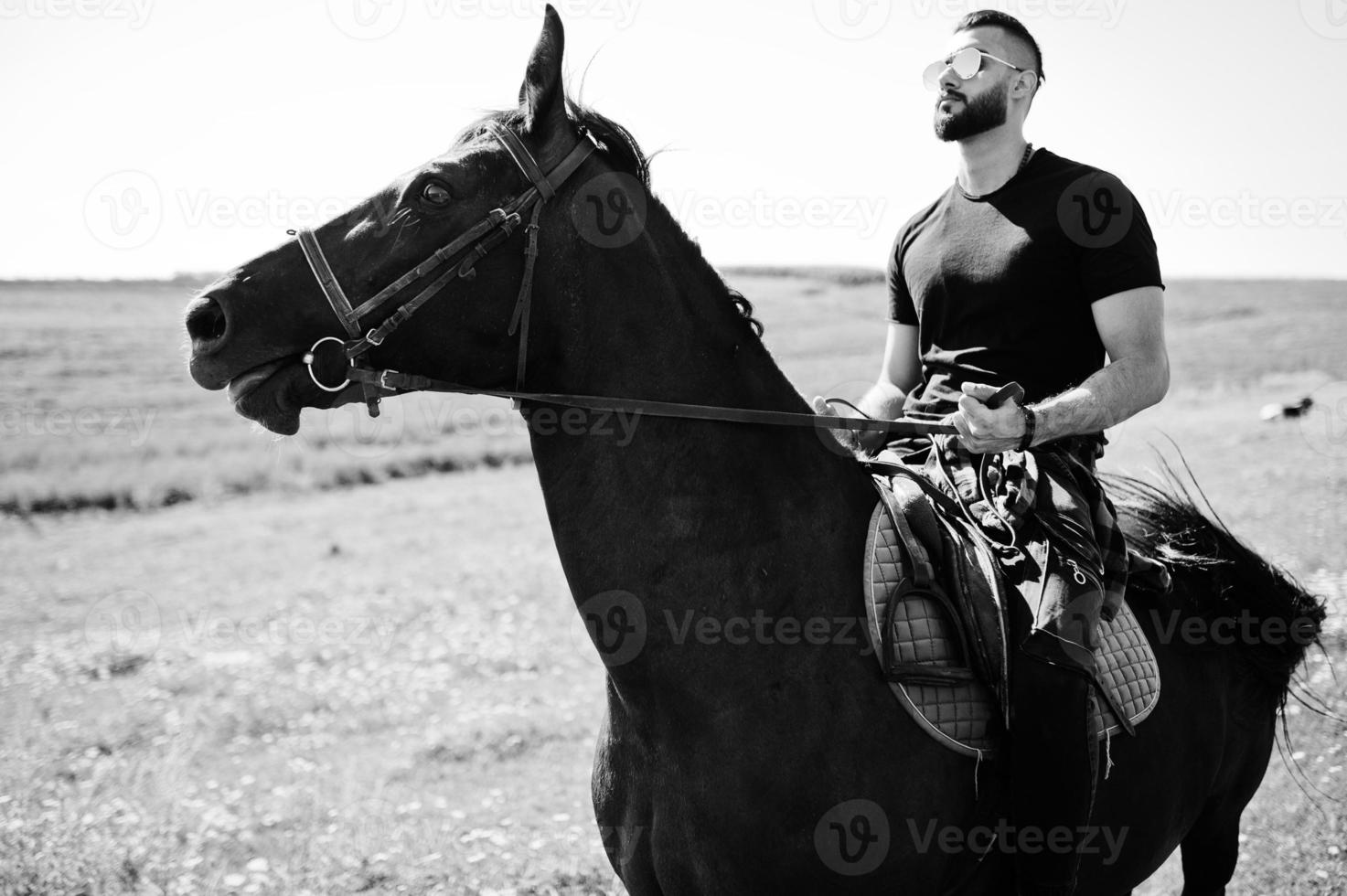 homem de barba alta árabe usa preto e óculos de sol montam cavalo árabe. foto