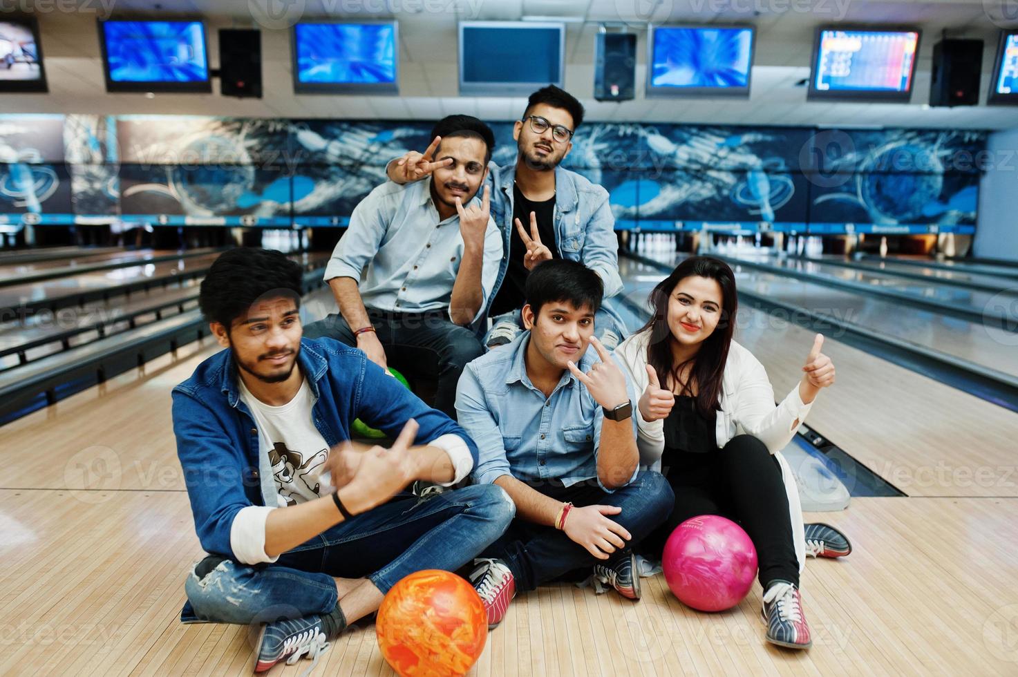 grupo de cinco povos do sul da Ásia descansando e se divertindo no clube de boliche, sentado na pista de boliche com bolas nas mãos. foto