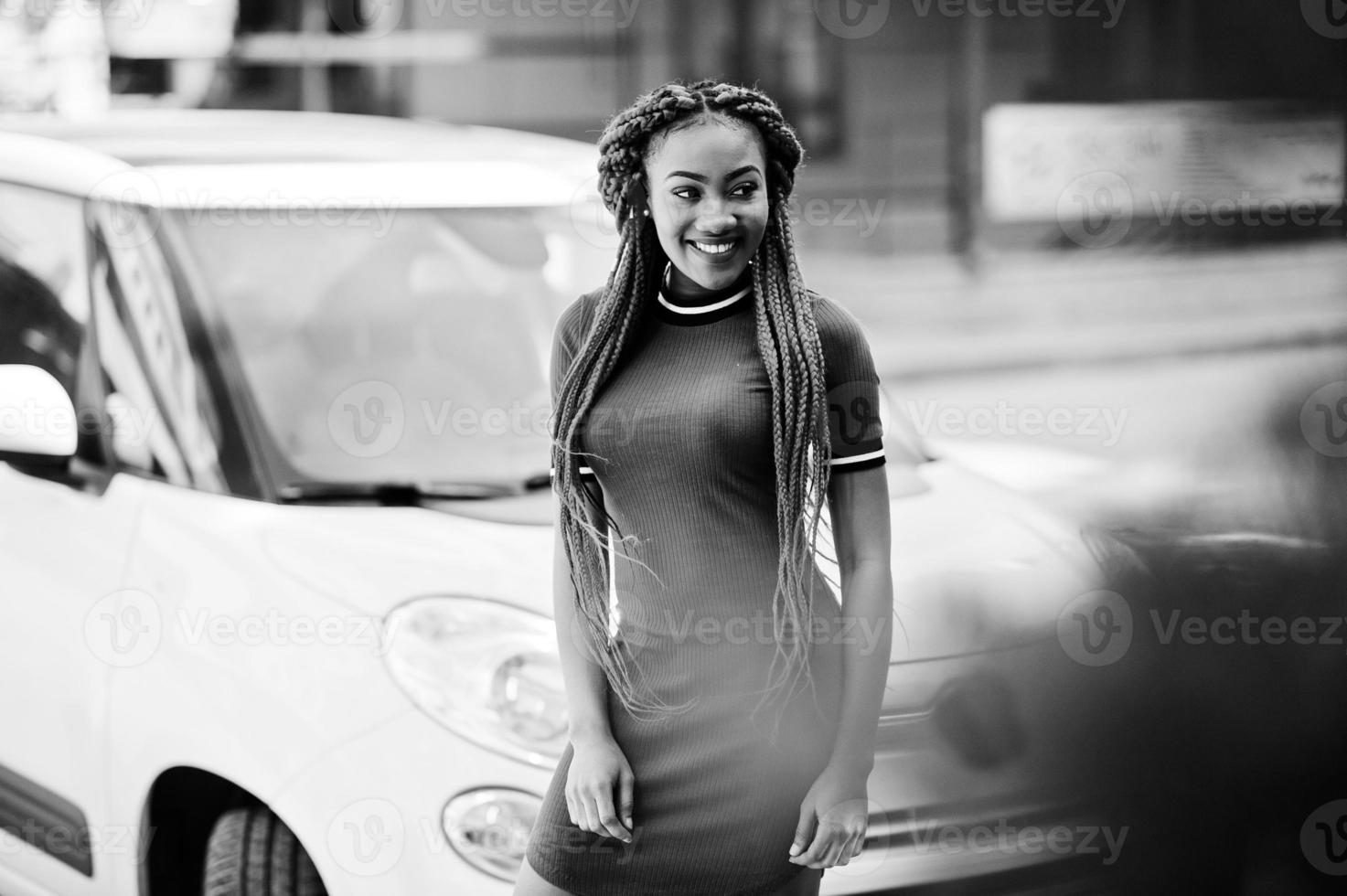 linda e magra garota afro-americana de vestido vermelho com dreadlocks posou ao ar livre no carro branco pequeno de fundo de rua. elegante modelo preto. foto