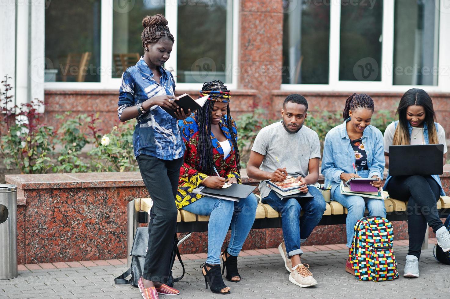 grupo de cinco estudantes universitários africanos passando tempo juntos no campus no pátio da universidade. amigos afro negros estudando no banco com itens escolares, notebooks laptops. foto