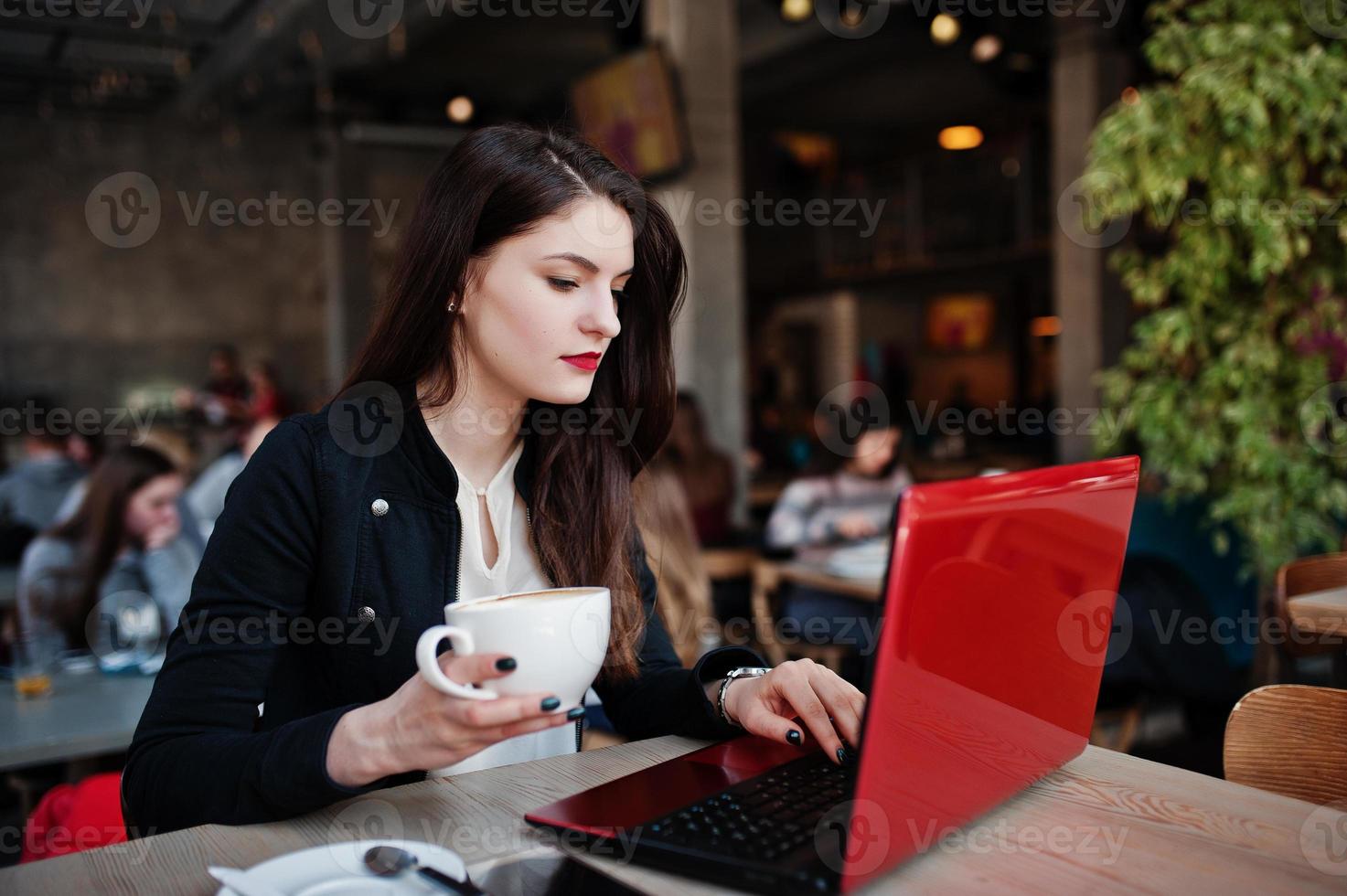 menina morena sentada no café com uma xícara de cappuccino, trabalhando com laptop vermelho. foto