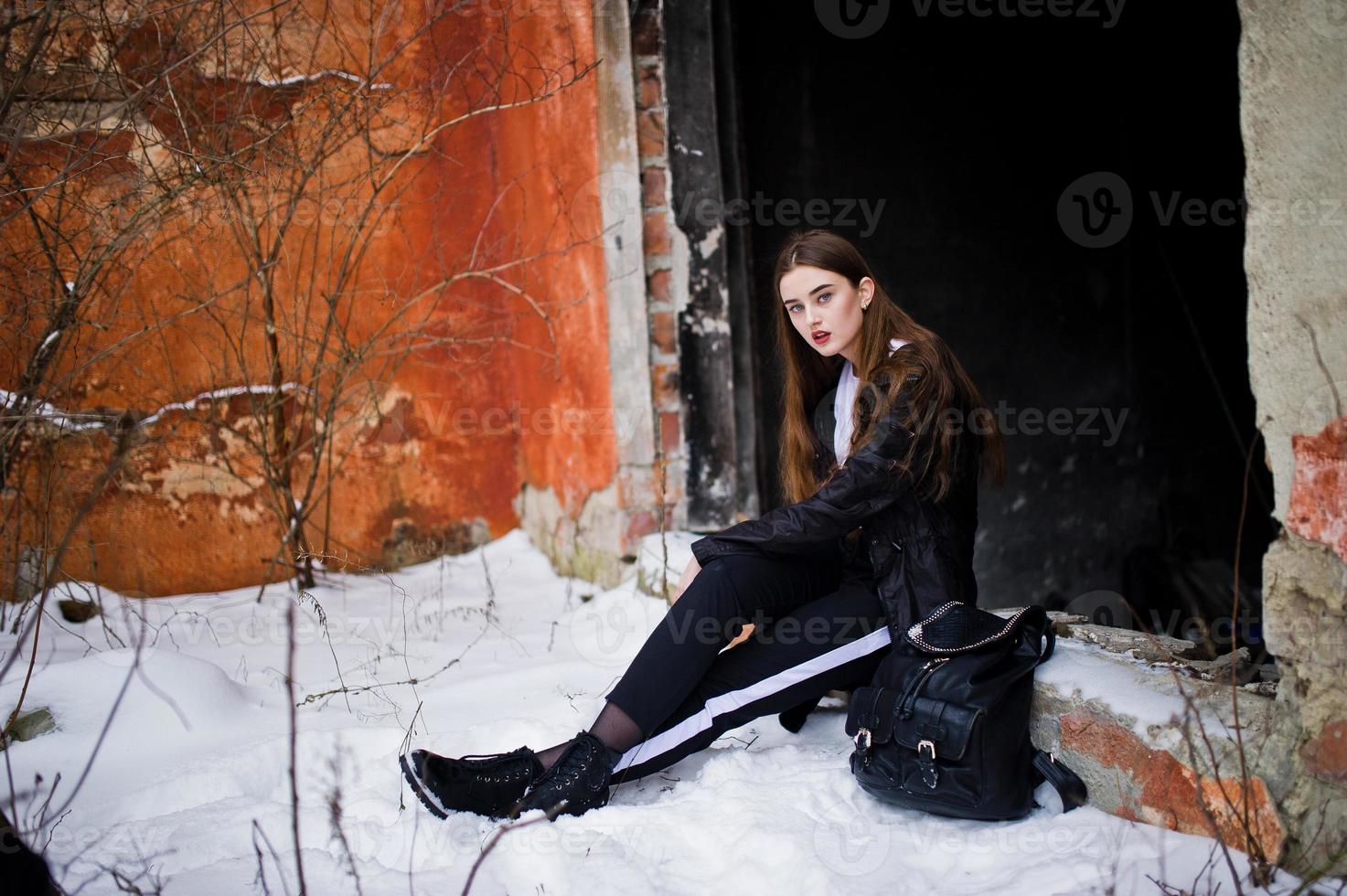 modelo morena de pernas longas na moda no manto preto longo posou ao ar livre no dia de inverno contra a velha parede do grunge. foto