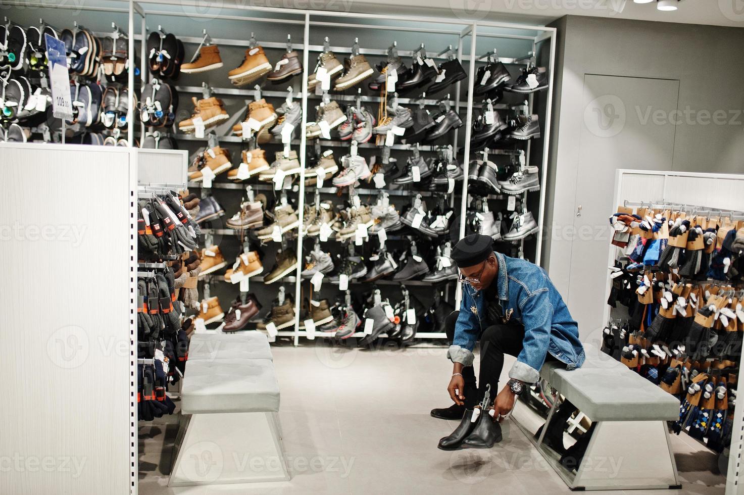 homem afro-americano casual elegante na jaqueta jeans e boina preta na loja de roupas tentando novos calçados. foto