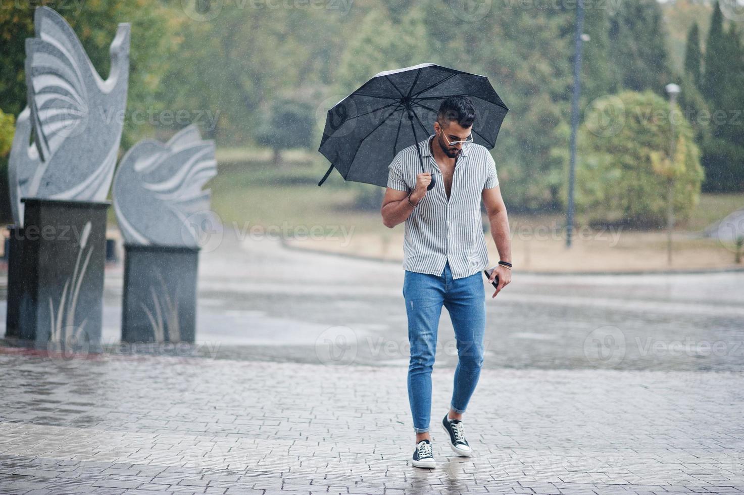 homem de barba árabe alto na moda usar camisa, jeans e óculos de sol com guarda-chuva posou na chuva na praça do parque. foto