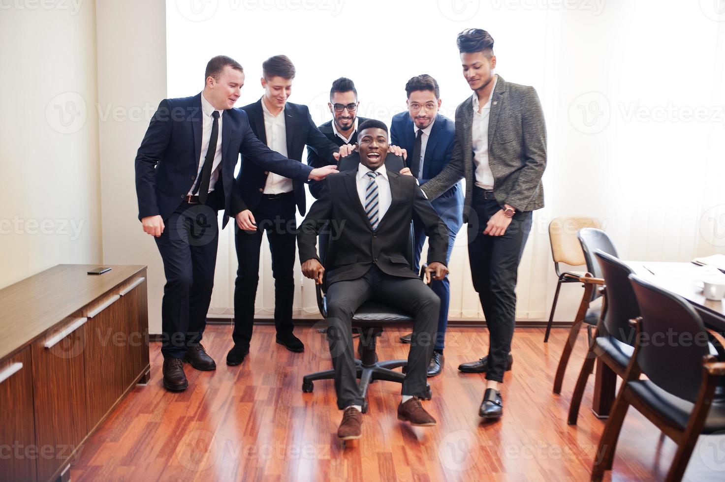 seis homens de negócios multirraciais em pé no escritório e rolar o homem na cadeira. grupo diversificado de funcionários do sexo masculino com roupa formal se divertindo. foto