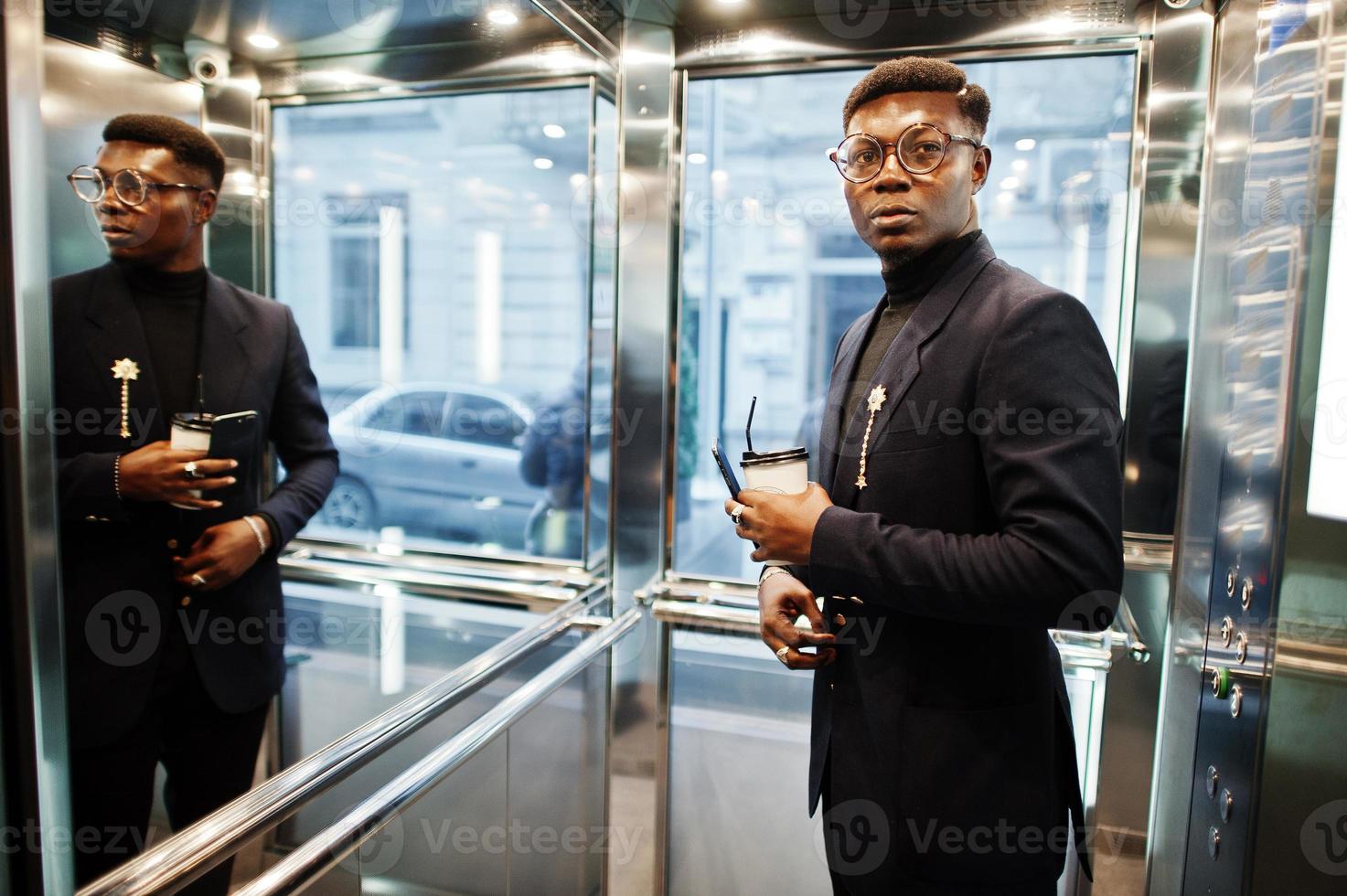 homem afro-americano elegante de terno e óculos com telefone celular e café nas mãos posou dentro do elevador. foto