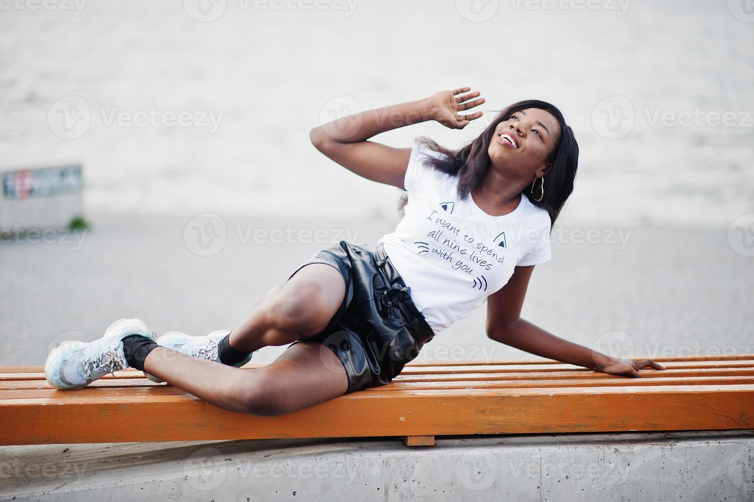 modelo magro de pele escura afro-americana posou em um short de couro preto e camiseta branca. ela luying no banco. foto