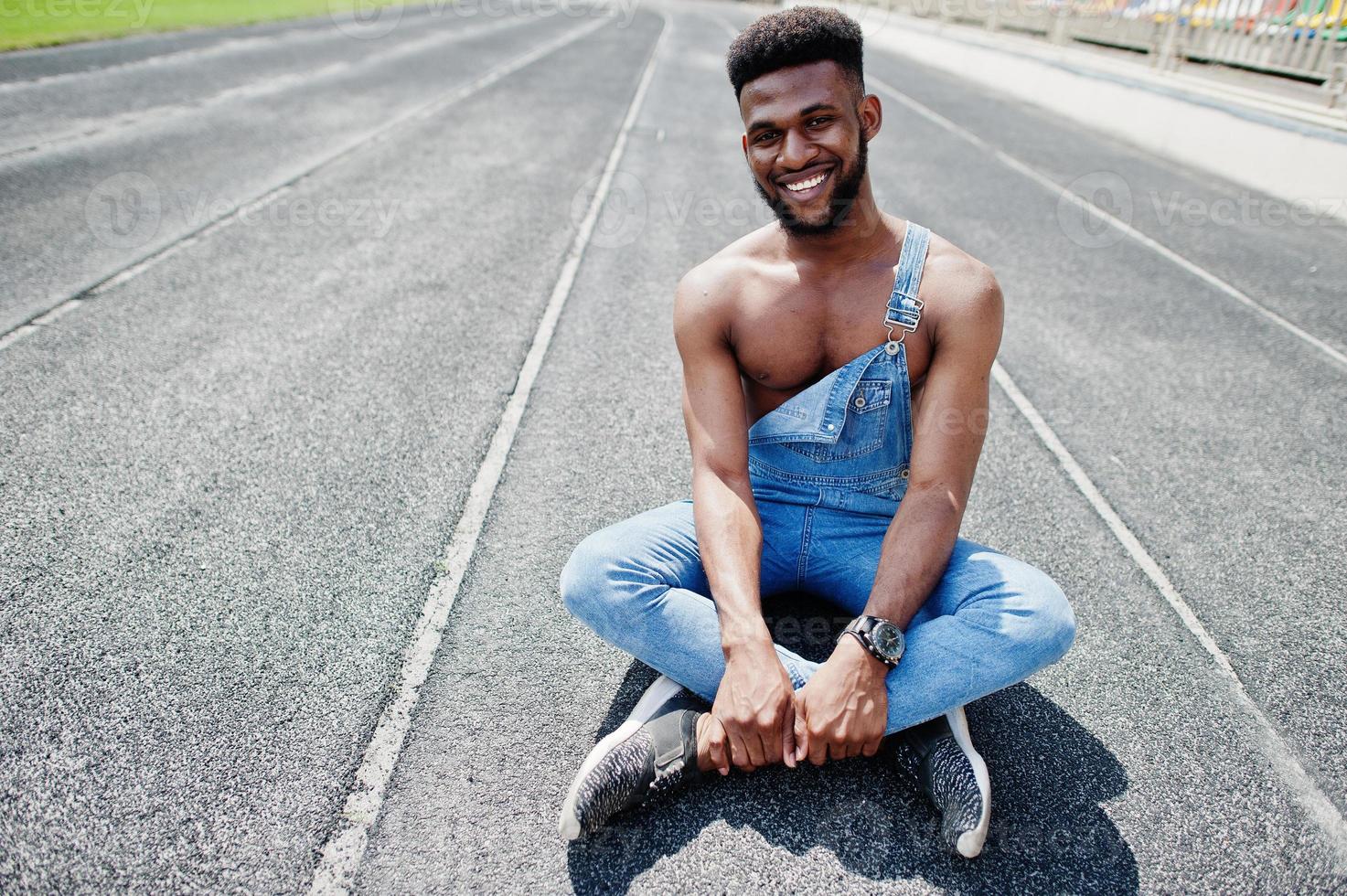 homem de torso nu afro-americano sexy bonito no macacão jeans sentado no autódromo do estádio. retrato de homem negro na moda. foto